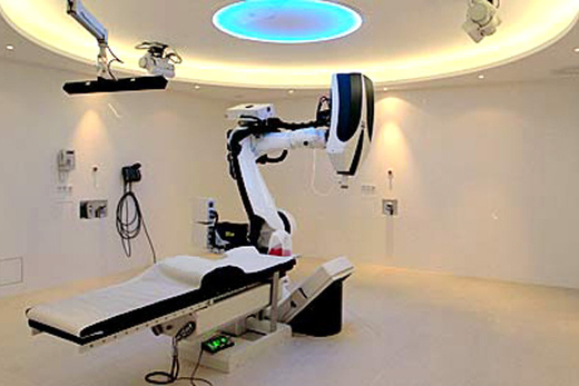 Behandlungsraum des Cyber-Knife-Zentrums München: Behandlungstisch, Roboter mit Linearbeschleuniger, an der Raumdecke die zwei Röntgengeräte des Bildortungssystems.